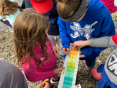 Children gather around a color-coded measurement chart by the water, engaged in a hands-on activity.