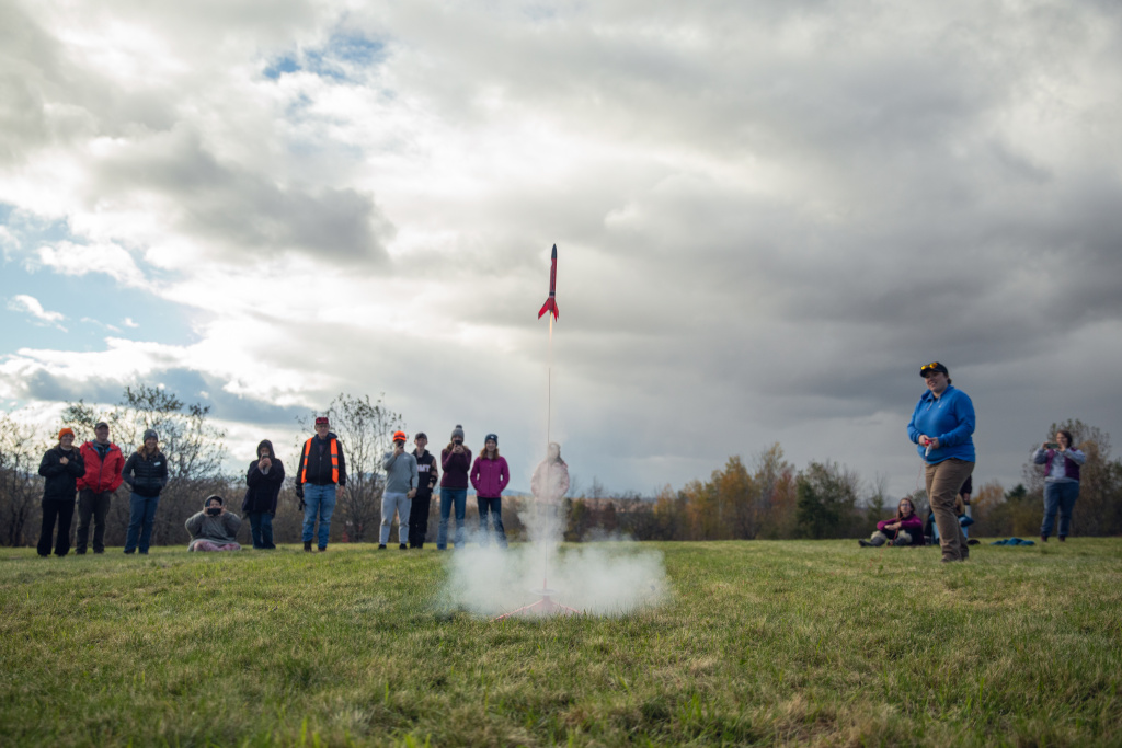 A group watches as a rocket launches into the sky, surrounded by clouds and grassy fields. Smoke billows from the launchpad.