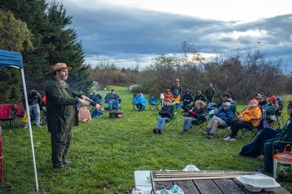 A speaker addresses a seated audience in a grassy area, surrounded by trees under an overcast sky.