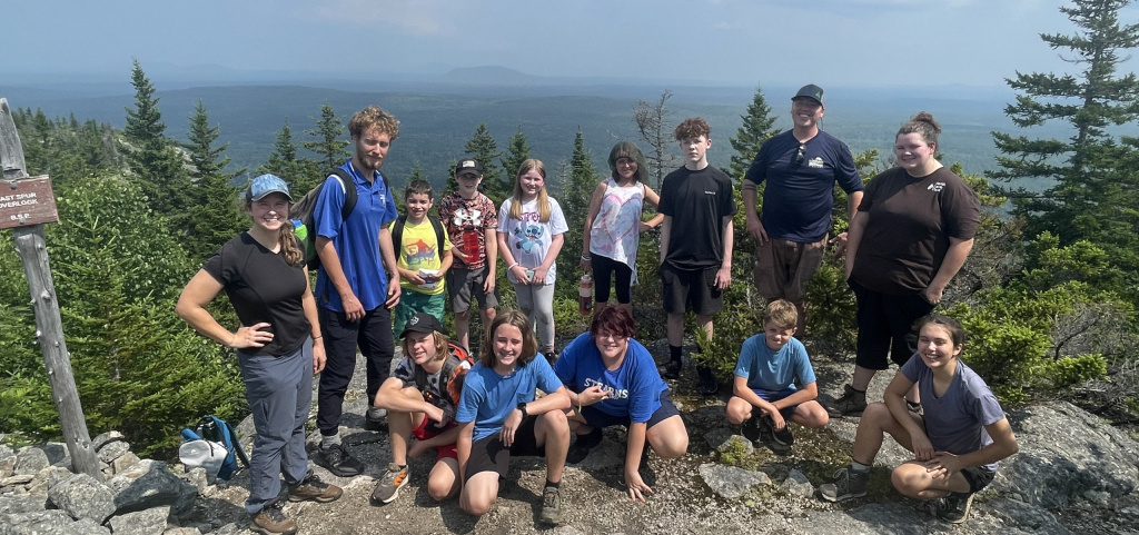 A group of adults and kids poses on a mountain overlook with green trees and a scenic view in the background.