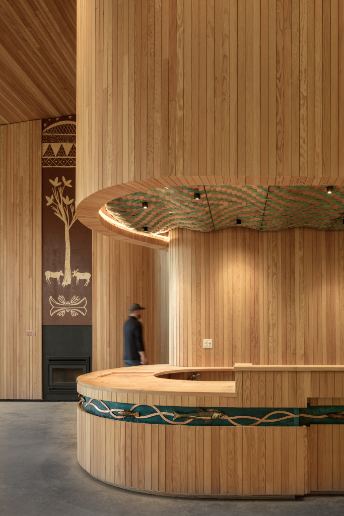 Wooden interior with curved desk and patterned ceiling; a person walks past artistic wall designs.