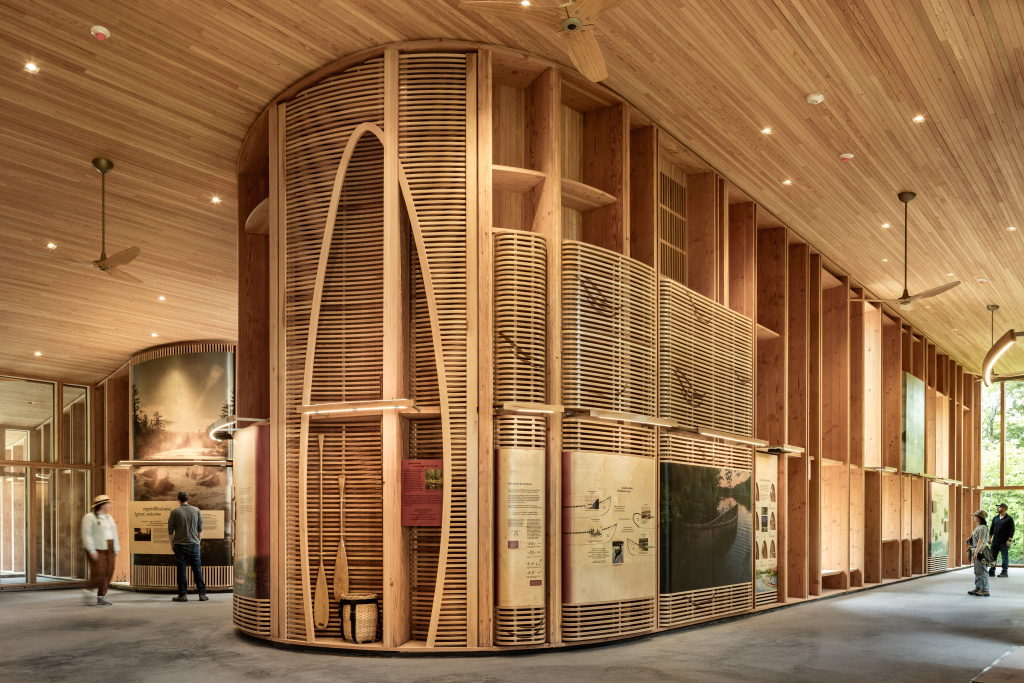 Interior of a wooden space with curved walls, art displays, and visitors exploring the area. Warm, natural light illuminates.