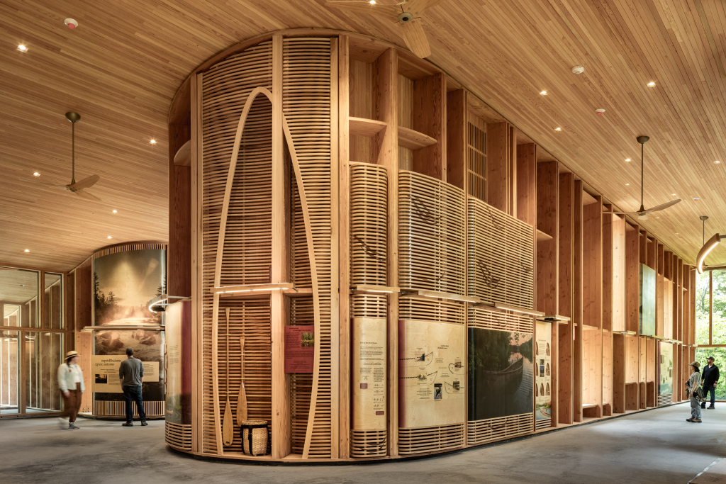 Modern wooden interior featuring curved walls, art displays, and visitors exploring an exhibit space.