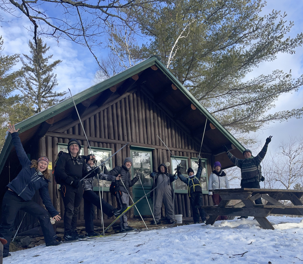 A group of happy people poses with ski poles in front of a rustic cabin, surrounded by snow and trees under a blue sky.