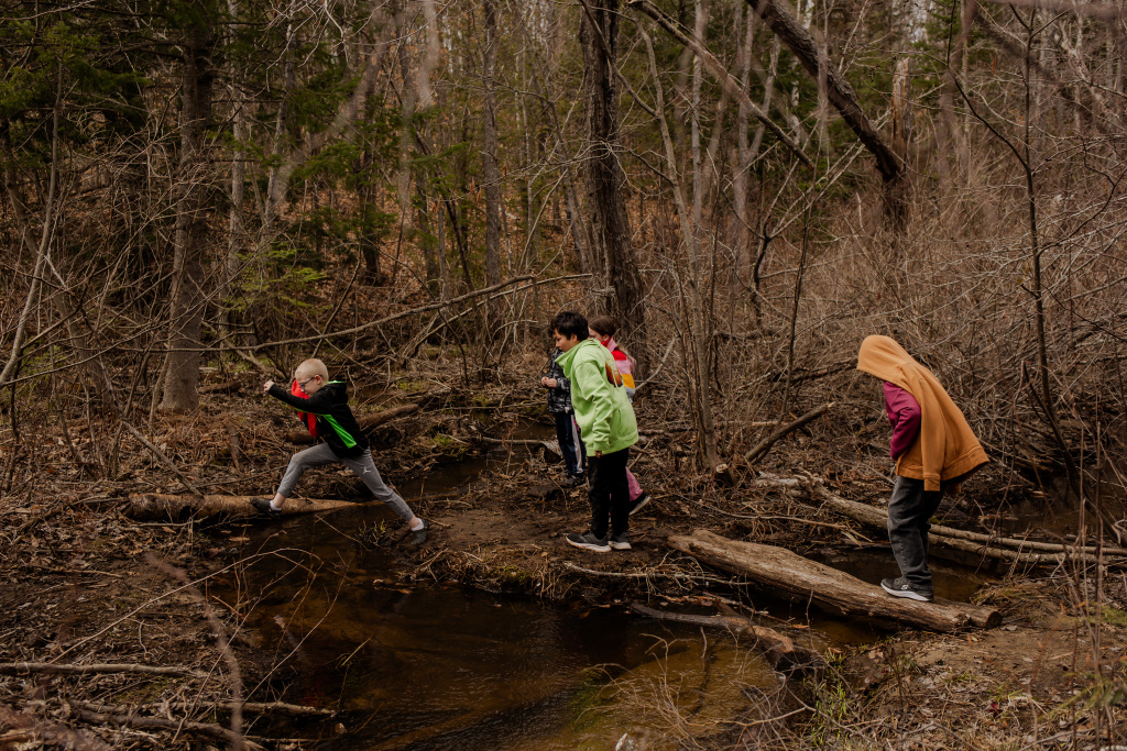 Children are exploring a wooded area, crossing a small creek on a log while dressed in colorful jackets.