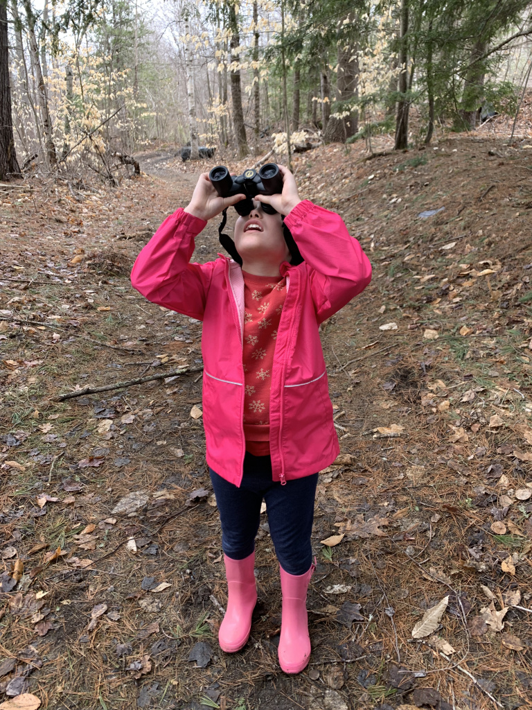 A child in a pink jacket and rain boots looks up with binoculars in a forested path.