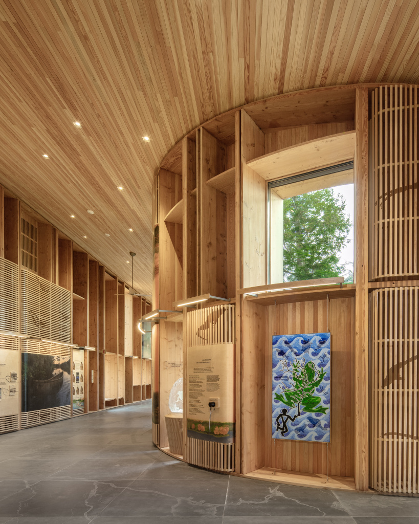 Interior of a modern wooden building featuring curved walls, art displays, and a large window with greenery outside.
