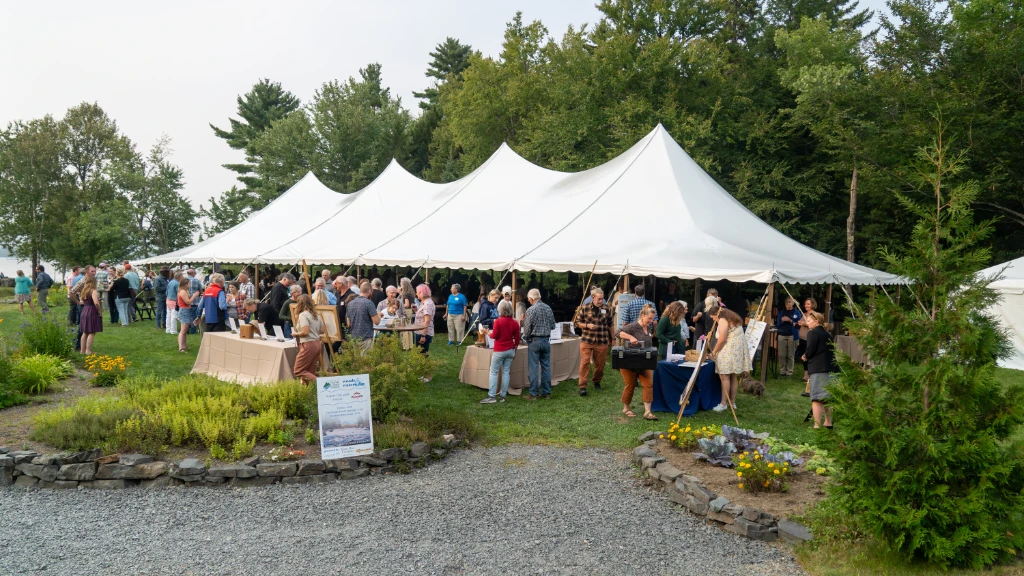 A lively outdoor gathering under a large tent with attendees mingling and enjoying the greenery.