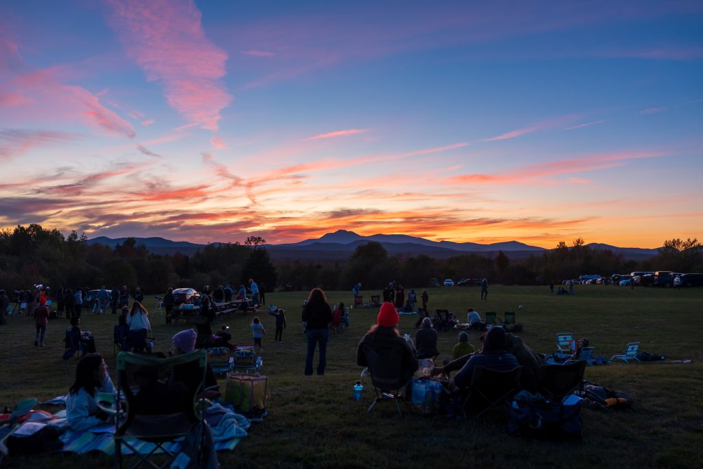 A group of people sitting on a grassy field, enjoying a vibrant sunset with mountains in the background.