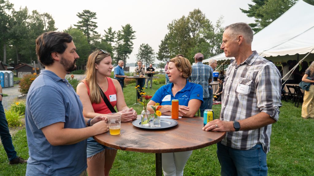 A group of four people chat around a table at an outdoor event, with drinks and flowers on the table.
