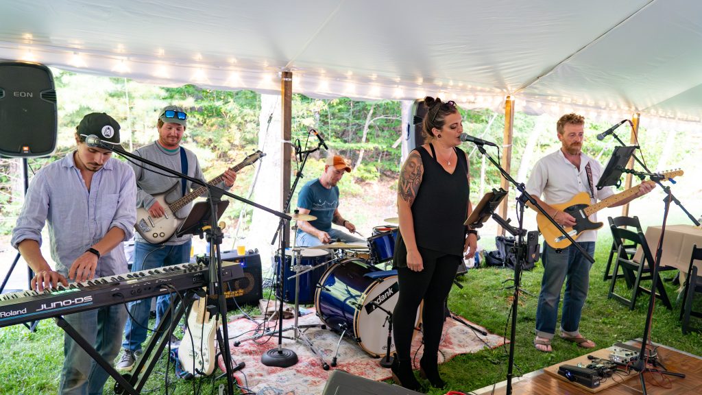 A band performs under a tent, featuring a singer, keyboardist, bassist, drummer, and guitarist. Greenery in the background.