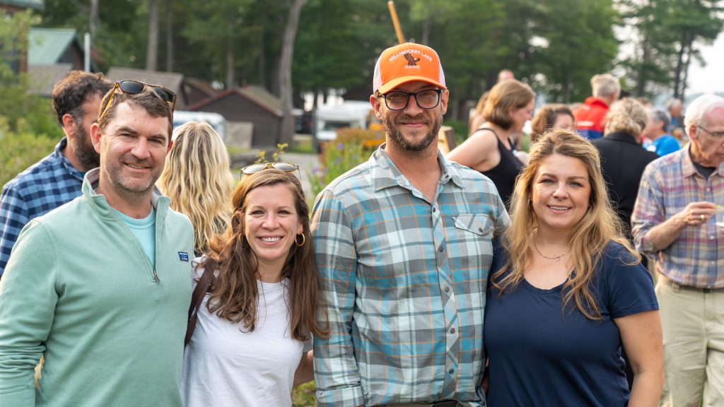Four people pose together outdoors, smiling amidst a crowd in a casual, sunny setting. Trees and buildings are in the background.