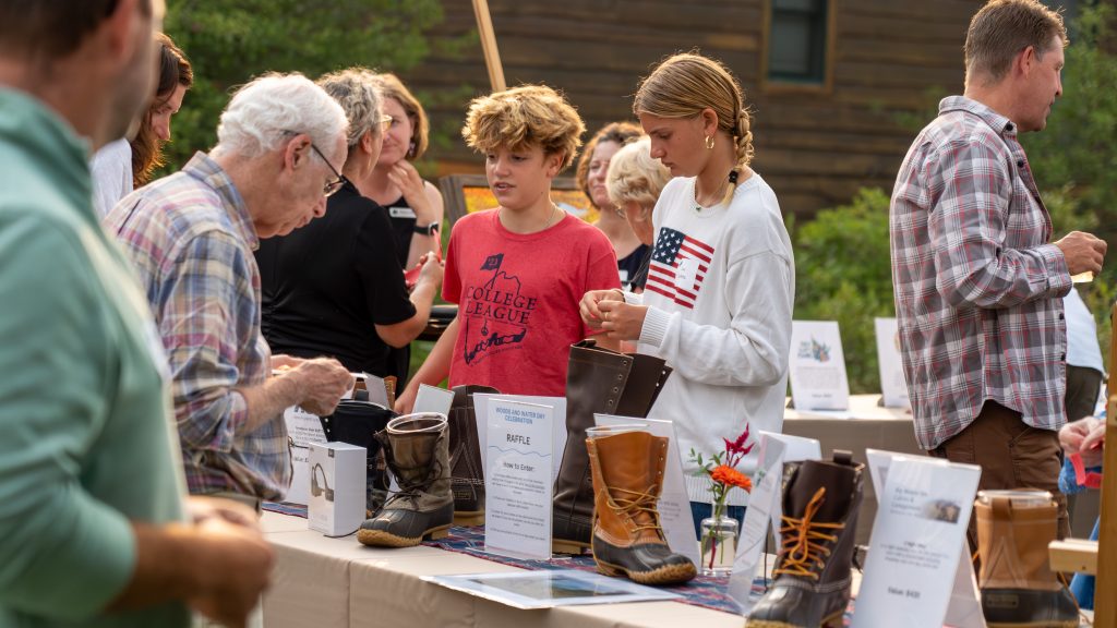 People gather at a table displaying various boots and raffle information during an outdoor event.
