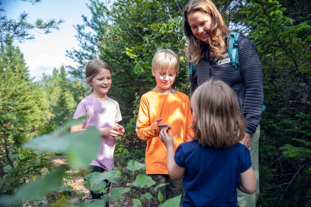 A woman and three children smile and interact while exploring a lush, green forest trail.