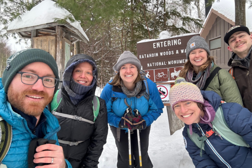 Six people are dressed in winter coats and standing outdoors in the snow, smiling at the camera.