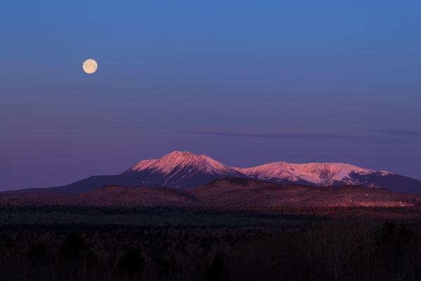 At twightlight, a full moon is high in the sky above a large snow covered mountain range.