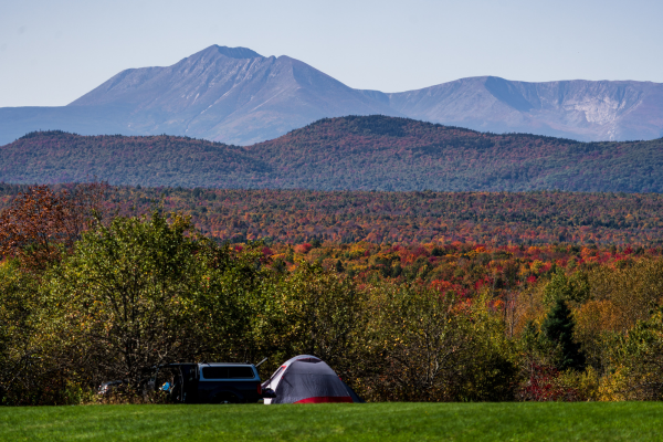 A large multi-peaked mountain rises in the disance behind a forested fall landscape, wiith a pickup truck and tent on a grassy field in the foreground.