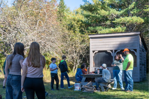 Teens are gathered casually around a camping pavillion and picnic table next to an old apple tree.