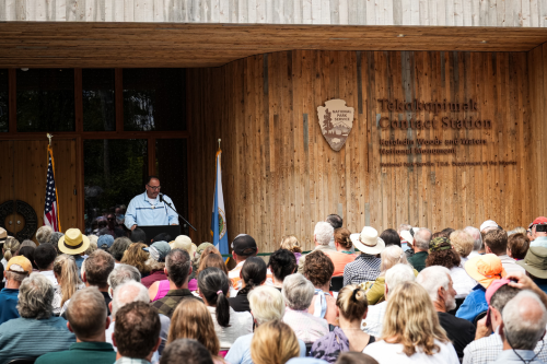 A man speaks from a podium at a gathering in front of Tekαkαpimək Contact Station.