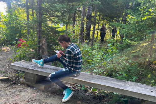 A teen wearing a plaid shirt and converse sneakers sits on a rustic wood bench in the woods sketching.