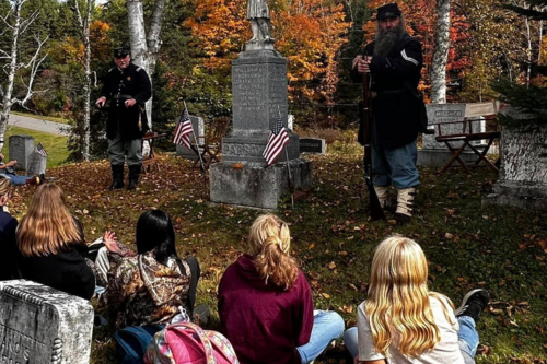 Four teen girls are seated on the ground in a cemetery watching two men in Civil War attire next to a memorial stone.